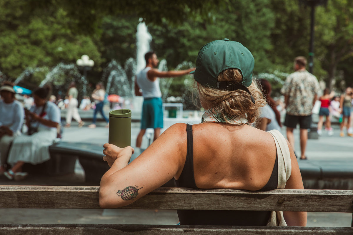 A woman in a cap sits at a park fountain with a coffee in her hand